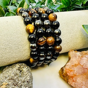 May include: A set of three beaded bracelets featuring black, brown, and silver beads. The bracelets are stacked on a white surface.