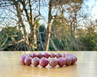 Puede incluir: Un brazalete de cuentas con piedras preciosas rosas redondas y pulidas. El brazalete descansa sobre una superficie marrón claro, con un fondo borroso de árboles. Las piedras preciosas tienen un aspecto natural y terroso.