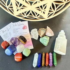 Könnte beinhalten: Eine Sammlung farbiger Kristalle und Edelsteine, darunter Rosenquarz und Amethyst, zusammen mit Informationskarten. Im Hintergrund befindet sich ein dekoratives Holzstück mit Schnitzereien. Die Kristalle variieren in Größe und Form.