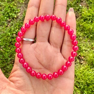 Puede incluir: Una pulsera de cuentas rosa brillante. La pulsera está compuesta por pequeñas cuentas redondas, creando un bucle continuo. Las cuentas son de un color rosa brillante y saturado. La pulsera está en la mano de una persona, con un anillo plateado visible.