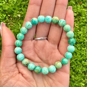 May include: A turquoise beaded bracelet with a silver ring. The bracelet features round, semi-precious stones in shades of turquoise and green. The bracelet is displayed on a hand with a green background.