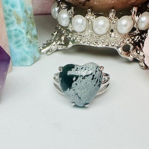 Puede incluir: Un anillo de plata con una piedra verde y blanca en forma de corazón. La piedra tiene un patrón moteado.