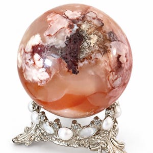 May include: A polished, spherical stone with translucent orange, white, and deep red inclusions, resting on a silver-toned metal stand decorated with small white pearls and clear crystals arranged in a crown-like circular pattern.