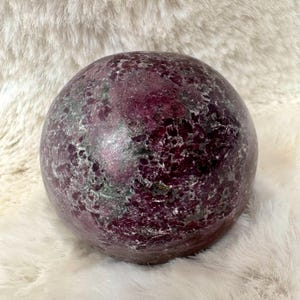 May include: A polished, spherical gemstone in shades of deep burgundy and dark grey. The surface has a textured, mottled appearance. The sphere is displayed on a white, fluffy surface, creating a contrast in textures.