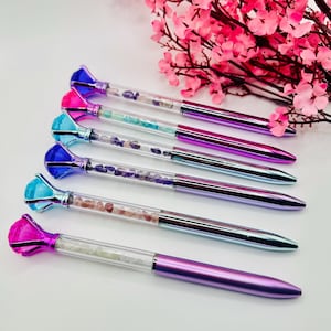 Op de afbeelding: Set van zes kleurrijke pennen met doorzichtige vaten gevuld met kleurrijke kristallen. De pennen hebben metallic paarse en zilveren vaten en diamantvormige toppen.