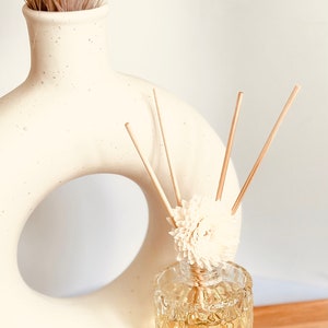 May include: A clear glass diffuser bottle with a textured surface and a white flower on top. The bottle is filled with a light yellow liquid and has wooden sticks sticking out of it. The diffuser is sitting on a wooden surface next to a white ceramic vase with a hole in the middle.