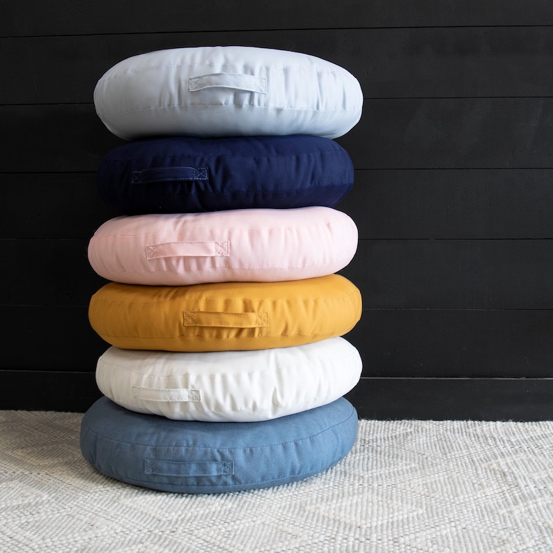 K&ouml;nnte beinhalten: Ein Stapel aus sechs runden Bodenkissen in verschiedenen Farben, darunter hellblau, dunkelblau, rosa, gelb, wei&szlig; und Jeansblau. Die Kissen sind in Pyramidenform angeordnet, wobei das gr&ouml;&szlig;te Kissen unten und das kleinste Kissen oben liegt.