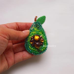 Pode incluir: Broche de abacate com contas em tons de verde, com um centro de gemas castanhas e laranja. O broche tem uma pequena folha verde e &eacute; segurado em uma m&atilde;o. O texto "Bzzjewelry" &eacute; vis&iacute;vel no lado direito.