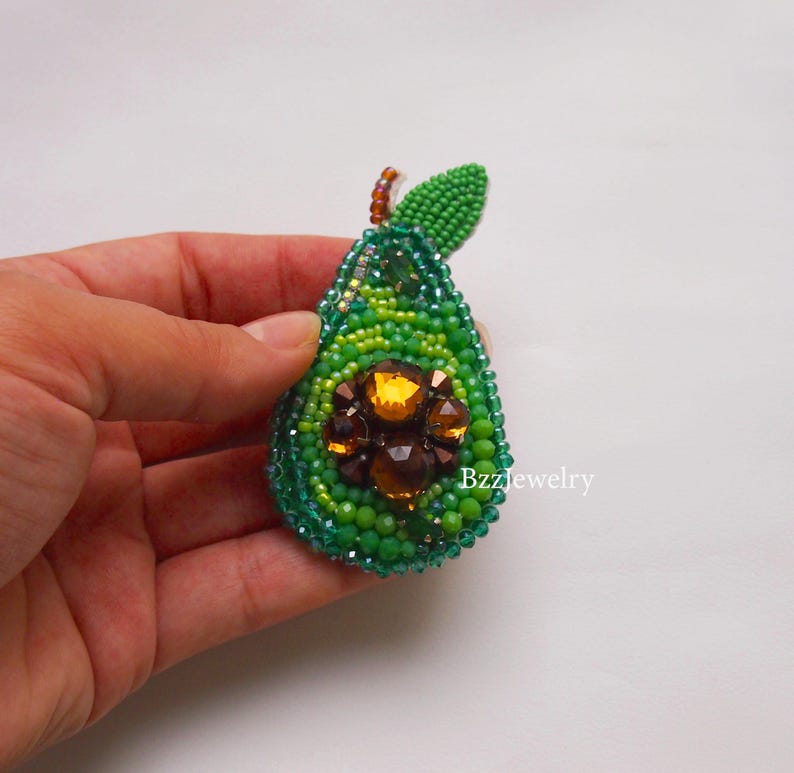 Pode incluir: Broche de abacate com contas em tons de verde, com um centro de gemas castanhas e laranja. O broche tem uma pequena folha verde e &eacute; segurado em uma m&atilde;o. O texto "Bzzjewelry" &eacute; vis&iacute;vel no lado direito.