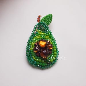 Pode incluir: Broche de abacate com contas em tons de verde, com um centro castanho e laranja. O broche tem um detalhe de folha no topo e &eacute; feito com pequenas contas. O design &eacute; intrincado e detalhado, com um acabamento brilhante.