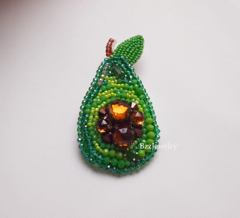 Pode incluir: Broche de abacate com contas em tons de verde, com um centro castanho e laranja. O broche tem um detalhe de folha no topo e &eacute; feito com pequenas contas. O design &eacute; intrincado e detalhado, com um acabamento brilhante.