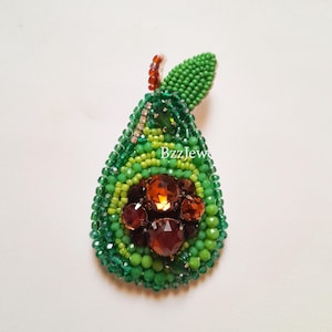 Pode incluir: Broche de abacate com contas em tons de verde e castanho. O broche apresenta um design em forma de p&ecirc;ra com um exterior de contas verdes, um centro de contas castanhas e uma pequena folha de contas verdes. O texto "BzzJew" &eacute; vis&iacute;vel.