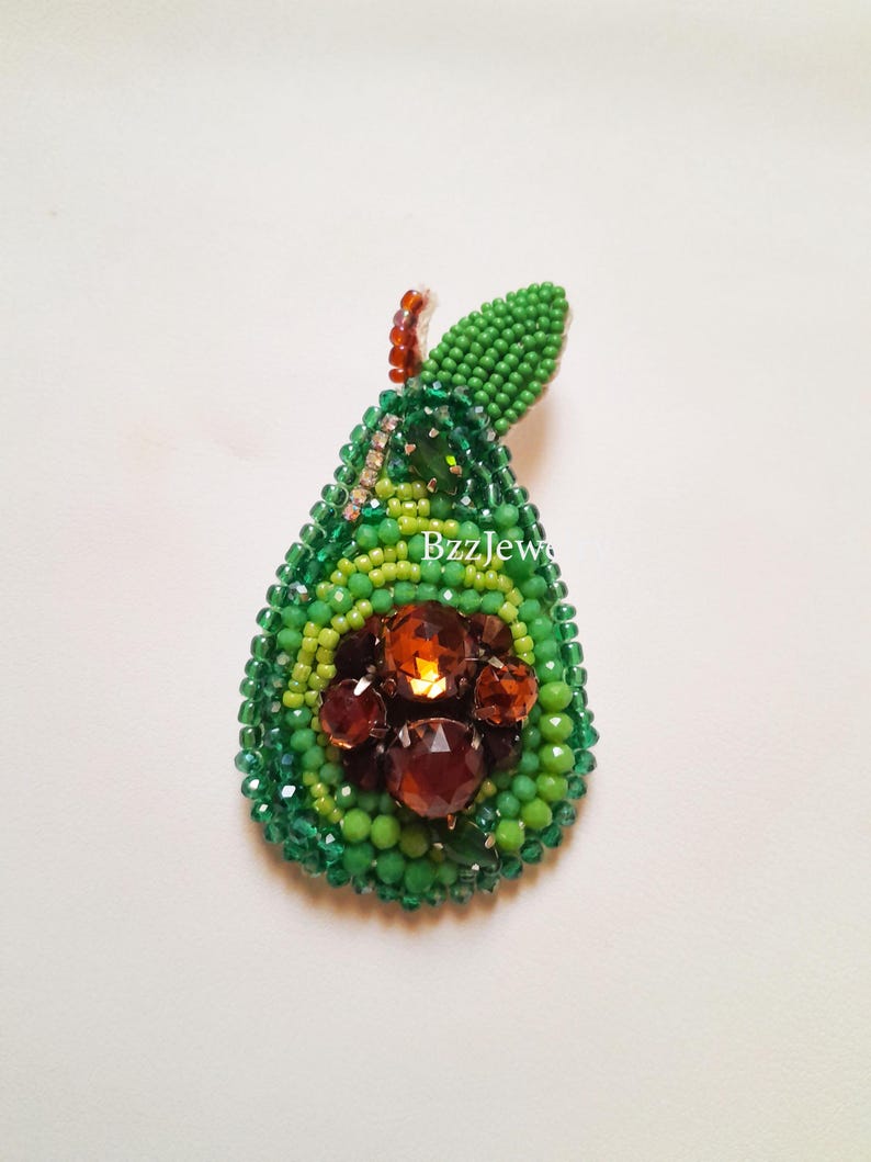 Pode incluir: Broche de abacate com contas em tons de verde e castanho. O broche apresenta um design em forma de p&ecirc;ra com um exterior de contas verdes, um centro de contas castanhas e uma pequena folha de contas verdes. O texto "BzzJew" &eacute; vis&iacute;vel.