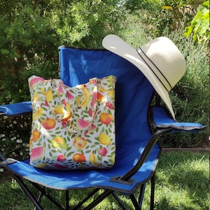 May include: A white canvas tote bag with a colorful fruit print design. The bag is sitting on a blue folding chair with a white sun hat on top of the chair.