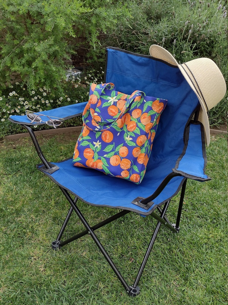 May include: A blue folding chair with a tote bag featuring a pattern of oranges and white flowers. The chair is sitting on green grass.