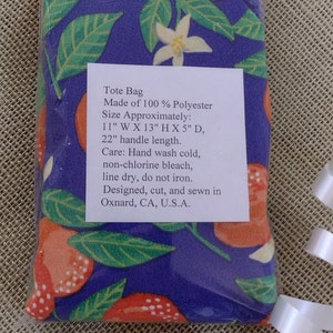 May include: A tote bag made of 100% polyester with a floral print of orange blossoms and red berries on a purple background. The bag measures 11 inches wide by 13 inches high by 5 inches deep, with a 22 inch handle. Care instructions include hand washing cold, non-chlorine bleach, line drying, and not ironing. The bag is designed, cut, and sewn in Oxnard, CA, U.S.A.
