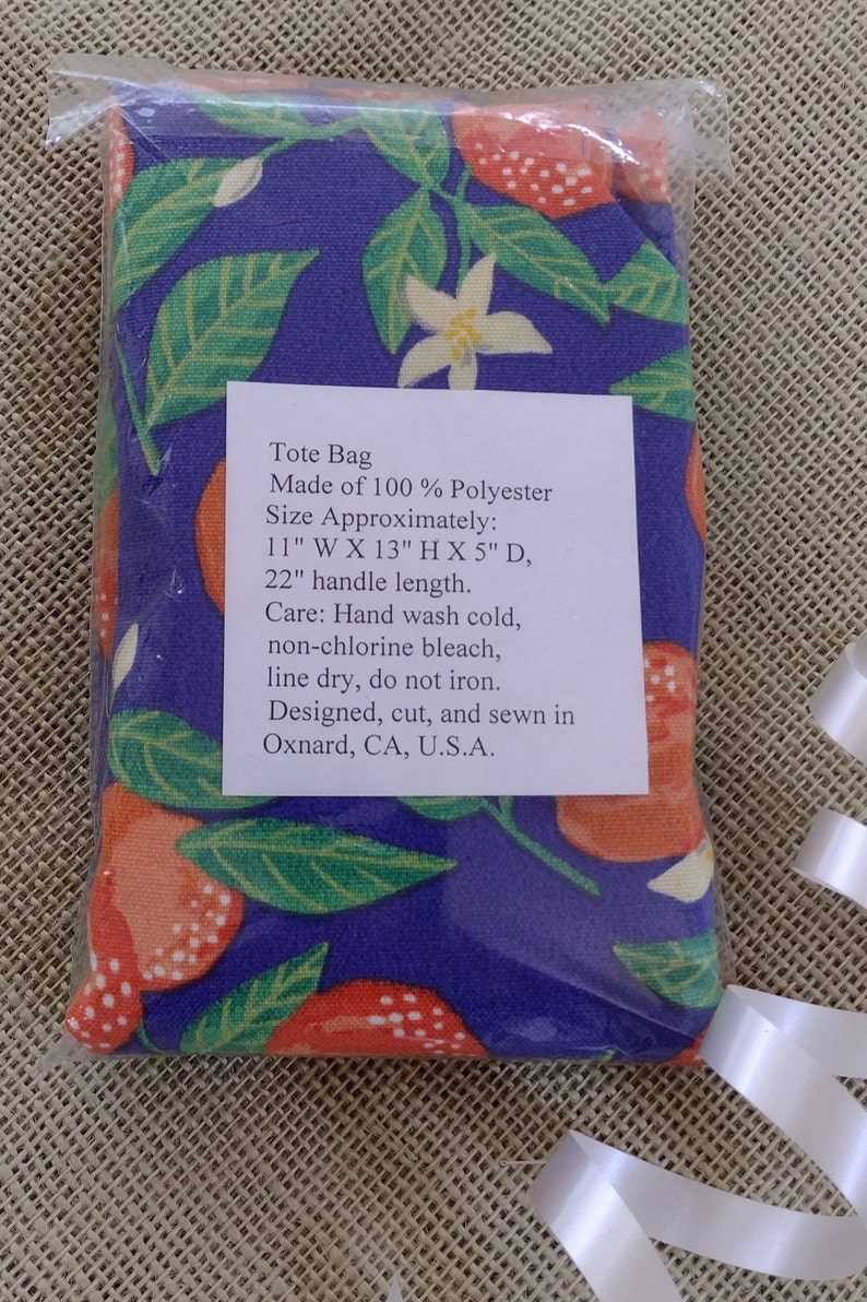 May include: A tote bag made of 100% polyester with a floral print of orange blossoms and red berries on a purple background. The bag measures 11 inches wide by 13 inches high by 5 inches deep, with a 22 inch handle. Care instructions include hand washing cold, non-chlorine bleach, line drying, and not ironing. The bag is designed, cut, and sewn in Oxnard, CA, U.S.A.