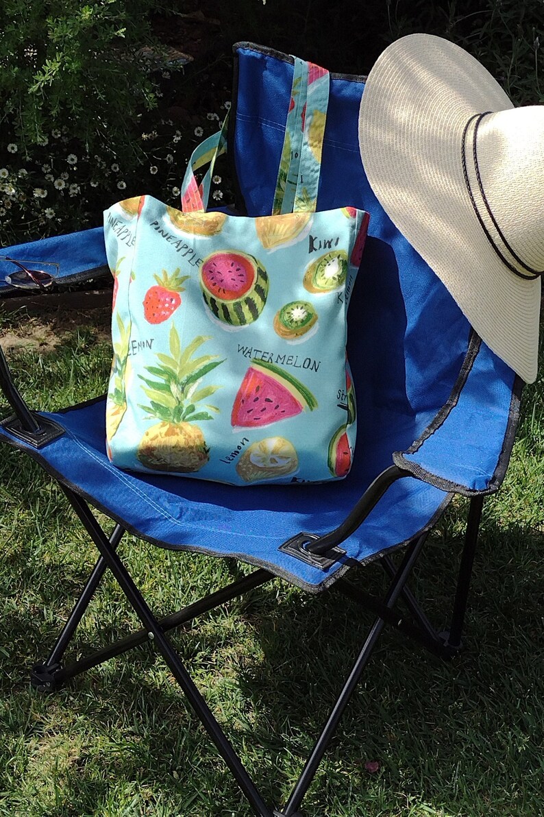 Peut inclure: Une chaise pliante bleue avec un sac fourre-tout imprim&eacute; de fruits. Le sac a un fond bleu clair avec des illustrations de past&egrave;que, de kiwi, d'ananas, de citron et de fraises. Le texte sur le sac est "Watermelon, Kiwi, Pineapple, Lemon".