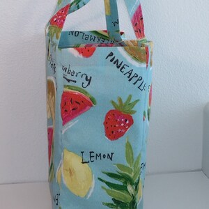 Peut inclure: Un sac cabas bleu clair avec un motif imprim&eacute; de fruits. Le sac pr&eacute;sente des fraises, des citrons, des past&egrave;ques et des ananas. Les mots "Ananas", "Citron" et "Fraise" sont imprim&eacute;s sur le sac.
