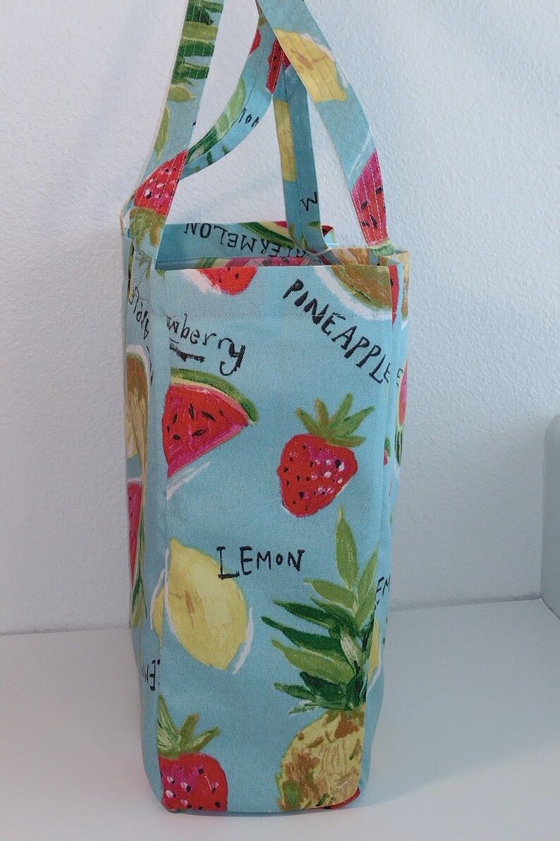 Peut inclure: Un sac cabas bleu clair avec un motif imprim&eacute; de fruits. Le sac pr&eacute;sente des fraises, des citrons, des past&egrave;ques et des ananas. Les mots "Ananas", "Citron" et "Fraise" sont imprim&eacute;s sur le sac.