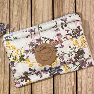 May include: A white quilted makeup bag with a floral print in shades of yellow, purple, and brown. The bag is tied with twine and has a brown tag that reads "Handmade Just for You".