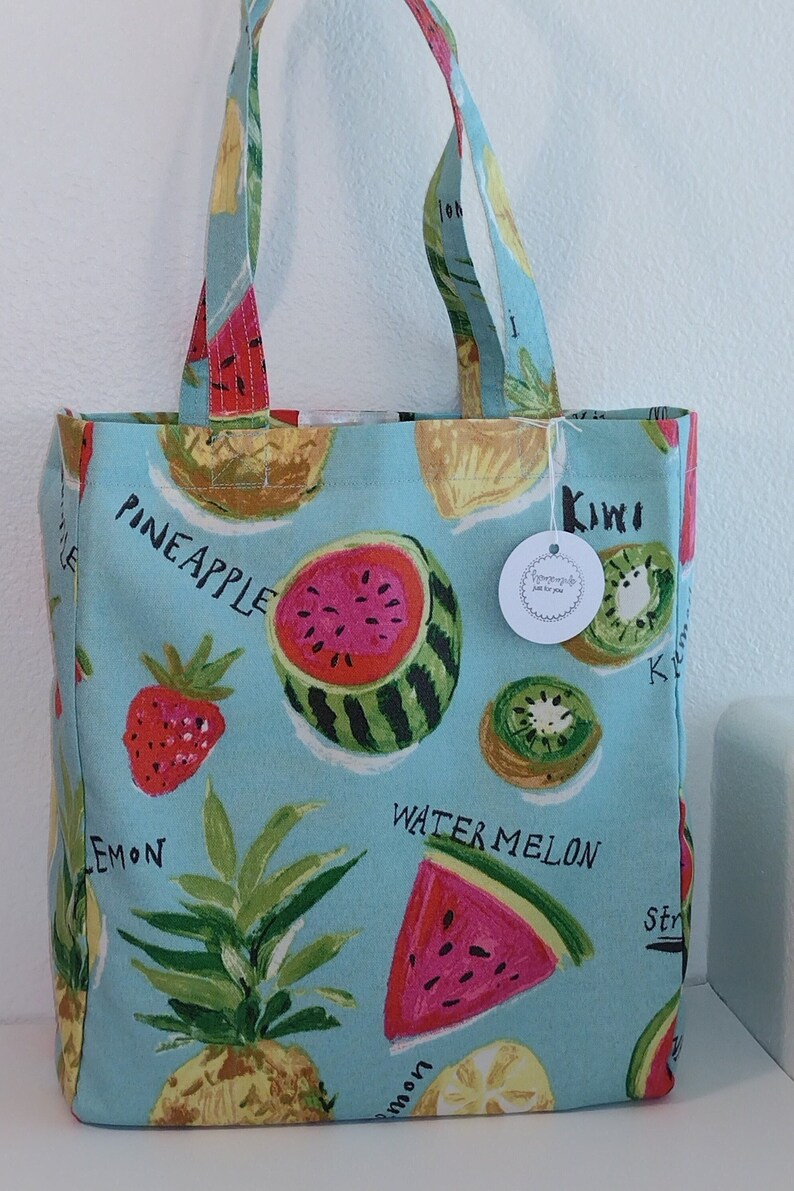 Peut inclure: Un sac cabas bleu clair avec un motif imprim&eacute; de fruits. Le sac pr&eacute;sente des past&egrave;ques, des ananas, des citrons, des fraises et des kiwis. Les mots "Pineapple", "Kiwi", "Watermelon", "Lemon" et "Strawberry" sont imprim&eacute;s sur le sac.