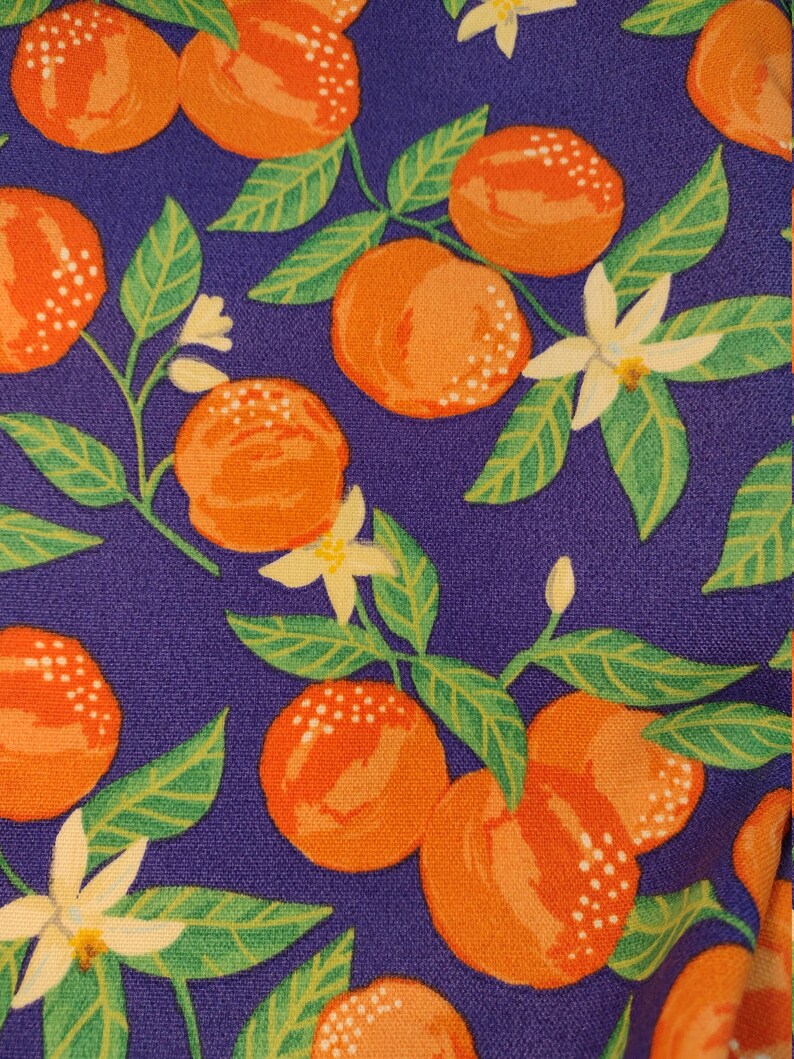 May include: Purple fabric with a repeating pattern of orange citrus fruit and white flowers. The fruit and flowers are on green stems and leaves.