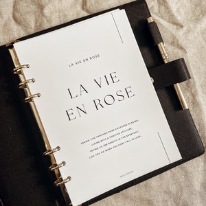 May include: A white planner with a black binder and gold rings. The cover features the text "La Vie En Rose" in a large, bold font. The text "Seeing life through rose-colored glasses. Living with a positive attitude. Trying to see beauty in the everyday. Like you do when you first fall in love." is printed below the title. The planner is made by Myla Paper.