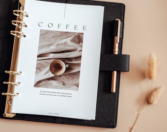Pode incluir: Uma agenda preta com uma encadernação em espiral dourada e uma página branca com a palavra "COFFEE" em letras grandes. A página também tem uma foto de uma xícara de café em um tecido branco. O texto abaixo da foto diz "Um líquido que cheira a café moído fresco, o céu e te coloca de bom humor." O texto "MILA PAPER" está na parte inferior da página.