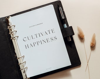 Może przedstawiać: Czarny segregator z otwartą białą stroną, na której widnieje tekst "Cultiver le bonheur" i "Cultivate Happiness". Na stronie znajduje się również tekst "Myla Fifer" na dole. Pióro i dwa suszone łodygi kwiatów leżą na stole obok segregatora.