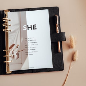 Könnte beinhalten: Ein schwarzer Planer mit einem goldenen Ringbinder und einer weißen Seite mit dem Text "SHE" und einer Liste positiver Affirmationen. Die Seite ist mit einem braunen Blattschatten verziert.