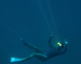 Pode incluir: Uma pessoa vestindo uma roupa de mergulho preta e uma máscara de mergulho amarela nada debaixo d'água com nadadeiras. A água é de um azul profundo e há um raio de luz brilhando de cima.
