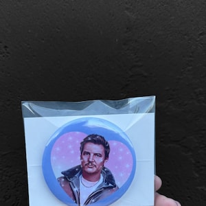 May include: A blue heart-shaped button with a man's face on it. The man has brown hair and a beard. He is wearing a blue jacket and a white shirt. The background is pink with white stars.