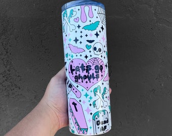 May include: A white tumbler with a pink and green design. The tumbler has a variety of spooky Halloween graphics, including bats, skulls, hearts, and crossbones. The text "Let's go ghouls" is printed on the tumbler.