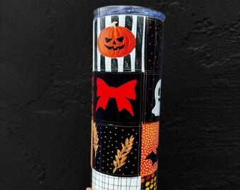 May include: A black and white tumbler with a patchwork design featuring Halloween-themed images, including a pumpkin, a bat, a ghost, and a bow. The tumbler is made of stainless steel and has a clear lid.