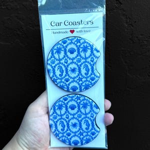 May include: Two blue and white car coasters with a tropical pattern of seahorses, shells, and palm trees. The coasters are packaged in a clear plastic sleeve. The packaging reads "Car Coasters Handmade with love."