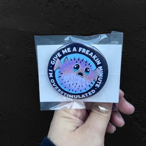 May include: A round button with a blue background and a white circle in the center. The white circle has the text "I'm Over Stimulated. Give Me A Freakin Minute." in black text. Inside the white circle is a cartoon illustration of a purple and pink pufferfish with black eyes and a sad expression. The pufferfish is surrounded by black spines.