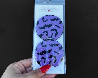 May include: Two purple car coasters with a black bat pattern. The coasters are packaged in a clear plastic sleeve with a white label that reads "Car Coasters Handmade with love".