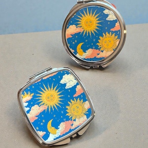 May include: Two silver compact mirrors, one round and one square, with celestial designs. The mirrors feature a blue background with yellow suns, a crescent moon, and pink and white clouds. The compacts are open, revealing the mirror surface.
