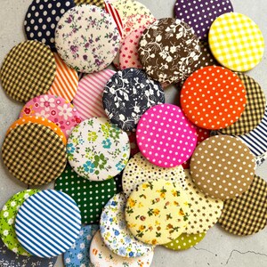 May include: A collection of circular fabric-covered buttons featuring a variety of patterns including polka dots, gingham checks, stripes, and floral prints in colors such as blue, yellow, pink, orange, green, brown, purple, and white.