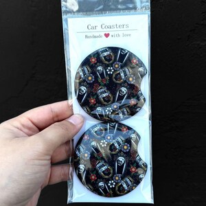 May include: Two black car coasters with a skull and crossbones design. The coasters feature a repeating pattern of skulls, crossbones, flowers, and the words "Rest in Peace". The text "Car Coasters" and "Handmade with love" is printed on the white packaging.