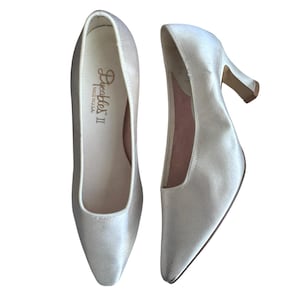 Könnte beinhalten: Ein Paar cremeweiße Satin-Pumps mit spitzer Zehenpartie und mittelhohem Absatz. Die Innensohle ist mit dem Schriftzug Dyeables II und MADE IN U.S.A. in Schreibschrift bedruckt. Die Schuhe haben eine glatte, glänzende Oberfläche und eine strukturierte Form.