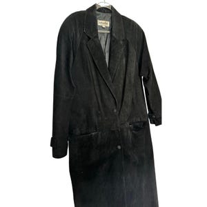 Puede incluir: Un abrigo largo de ante negro con un diseño cruzado. El abrigo presenta un cuello con muescas, dos bolsillos delanteros y cierres de botón. El abrigo está colgado de una percha de metal.