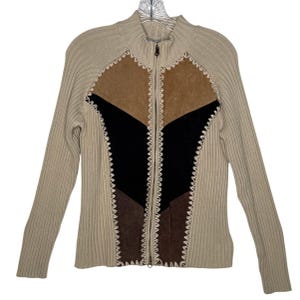 Peut inclure: Un pull beige zippé avec des manches et un corps en tricot côtelé. Le devant présente un motif géométrique en daim marron, noir et beige, cousu avec un motif décoratif en zigzag blanc. Le pull a un col montant.