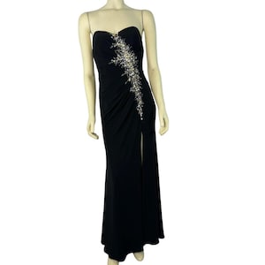 Puede incluir: Vestido de noche negro sin tirantes con una abertura alta. El corpiño está adornado con un diseño asimétrico de cristales plateados y transparentes. El vestido largo tiene una silueta ajustada.