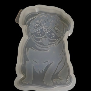 May include: A translucent silicone mold featuring a detailed recessed design of a sitting pug dog with its tongue sticking out. The mold has a raised outer border and a smooth, flexible surface, capturing the facial features and body shape of the dog.