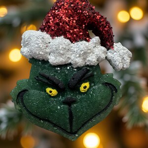 May include: A Christmas ornament featuring the Grinch's face, wearing a red sequined Santa hat with white glitter trim. The Grinch's face is green with yellow eyes and a black nose and mouth. The ornament is hanging from a silver hook.