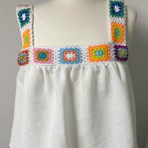 Pode incluir: Uma blusa branca sem mangas com decote quadrado adornado com uma fila de quadrados de crochê coloridos. As alças também são detalhadas com quadrados multicoloridos a condizer. O corpo principal da blusa é feito de tecido branco liso com um acabamento texturizado.