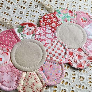 May include: Two flower-shaped quilted pot holders. Each pot holder is made of various pink and red floral fabrics. The centre of each flower is a beige circle. A beige lace loop is attached to the top of each pot holder.