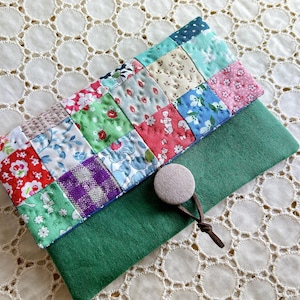 Puede incluir: Un bolso de mano de tela patchwork con base de fieltro verde. El patchwork presenta varias telas estampadas y florales en rojo, azul, verde y rosa. Se ve un botón beige y un cierre de cordón marrón.